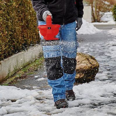 Person in blauer Arbeitskleidung verwendet einen roten Streuer auf einem schneebedeckten Gehweg.