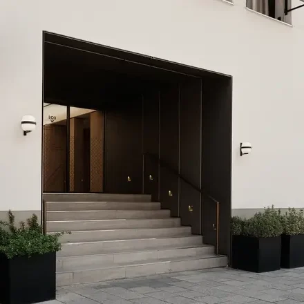 Eingang mit Treppe, beleuchtet von seitlichen Wandlampen und umgeben von Pflanzenkübeln.