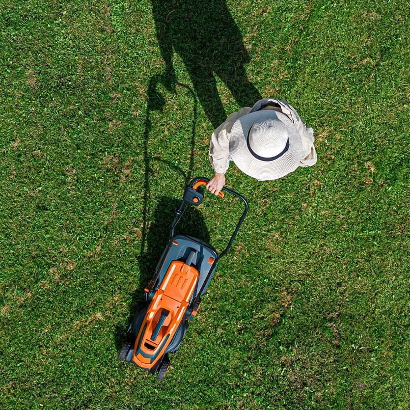 Person in einem Hut mäht Rasen mit einem elektrischen Rasenmäher.