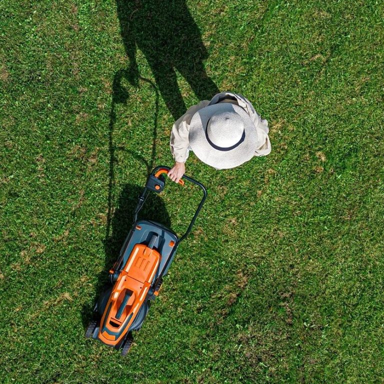 Person mit Hut mäht Rasen mit einem elektrischen Rasenmäher auf grünem Gras.